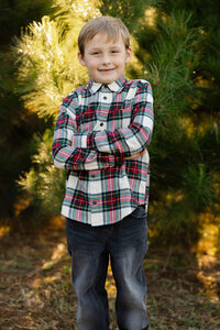 Young boy standing outdoors in front of evergreen trees, smiling in a plaid shirt during a fall family photo session by Kaitlyn Dawn Photography, children’s portrait photographer.