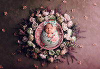 Newborn baby girl swaddled in a floral wrap and bow, sleeping peacefully in a pink basket surrounded by blush and lavender flowers, photographed in Waukesha near Hartland, WI.