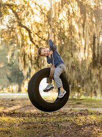 Young boy laughing as he hangs from a rope and rides a tire swing beneath moss-draped trees during an outdoor child portrait session. (Photographed by Nature's Child Photography)