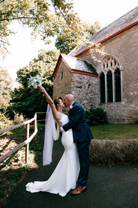 colourful and candid wedding photography at cockington court capturing bride and groom sharing a moment infont of church