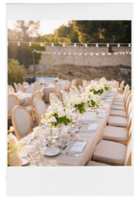 A long wedding reception table with white floral arrangements, cushioned chairs, and string lights overhead | Venue Business Boot Camp