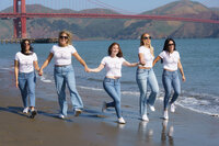 Five women skipping down the beach while dressed in white t-shirts and bluejeans in front of the golden gate bridge