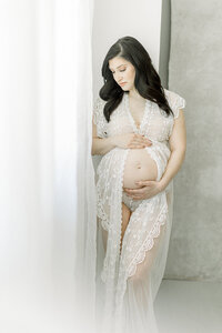 A pregnant mother dressed in a chiffon maternity gown while she is standing in front of a window looking down at her growing belly at a Dallas/Fort Worth photography studio.