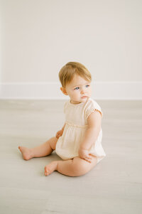 Baby girl in peach outfit sits and looks out window during photography studio in Raleigh NC