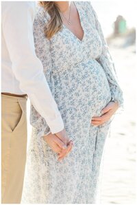A couple holds hands during maternity photo on the beachs. The wife is wearing a white dress with blue flowers, the husband is wearing a white shirt with khaki pants.