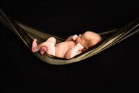 A naked baby lies in a soft green fabric against a black backdrop. The fabric against the black backdrop gives the illusion that the baby is lying in a hammock.