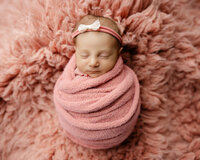 Sleeping newborn baby girl wrapped in a blush pink swaddle and posed on a soft textured blanket during a studio newborn photography session.