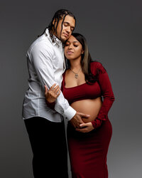 Romantic studio maternity portrait of an expecting couple embracing with mother wearing a fitted burgundy maternity dress while father gently holds her pregnant belly, captured by Kaitlyn Dawn Photography.