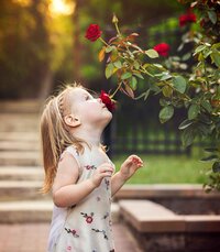 In Palm Coast, a wide-eyed little girl pauses to smell a bright red rose, her soft expression capturing the quiet magic of childhood during an outdoor. portrait session - A portrait experience by Nature's Child Photography.