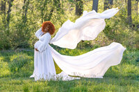 Pregnant woman in a flowing white gown with fabric billowing in the wind during a maternity photography session at Mill Creek Park