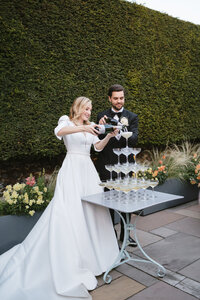 At an elegant outdoor wedding reception at the Chapel House Estate in Kent, a bride and groom, dressed in formal wedding attire, are lovingly pouring champagne into the top glass of a pyramid-shaped tower of empty glasses, creating a cascade of bubbles.