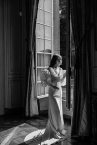 Black and white portrait of a bride in a long gown with sheer sleeves, standing by an open window and looking outside, softly lit by natural light.