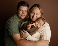 New parents smiling while holding their sleeping newborn baby wrapped in a soft brown swaddle during a studio newborn family photography session by Kaitlyn Dawn Photography.