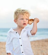Blonde haired boy holding a seashell up to his ear, listening to the oceans call in a child portrait session by Nature's Child Photography