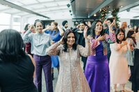 Women dancing at an indoor celebration, dressed in colorful attire, with others in the background also participating in the dance.