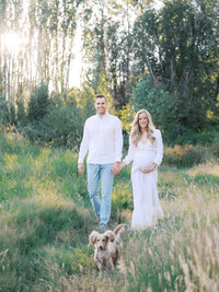 A couple dressed in white walking hand in hand with their dog running ahead during a maternity session in Magnuson park in Seattle