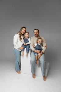 Mother standing beside father while holding their toddlers in a minimalist gray studio, candid family portrait by Kaitlyn Dawn Photography.