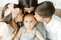 A family portrait of a mother and her kids sitting for an in studio photography session as they all lean in to kiss the baby boy.