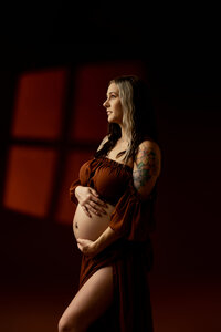 Pregnant woman in a rust-colored gown holding her baby bump with dramatic lighting during a Yucaipa studio pregnancy photography session with Kaitlyn Dawn Photography.