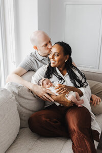 Husband kissing wife on cheek. She is holding newborn daughter photographed by Toronto Newborn Photographer Claire Binks