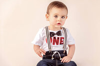 one year old boy in suspenders and bow tie sitting for milestone portrait in neutral studio by new bern family photographer