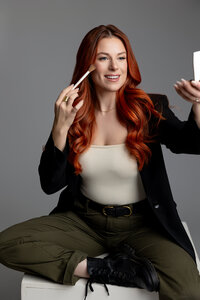 Woman with red hair seated at a desk applying mascara with compact mirror. Photograph by Yucaipa branding photographer Kaitlyn Dawn Photography.