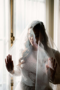 Emotive documentary storytelling wedding photography of a bride behind a sheer veil with her hands pressed against a window.