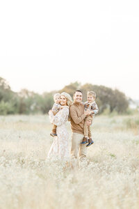 Beautiful family portrait of a mother and father holding their two young boys as they look and pose for family photos in the middle of an open field at a DFW park.