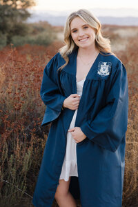 High school graduate wearing a cap and gown standing in a golden field during sunset, graduation portrait by Kaitlyn Dawn Photography.