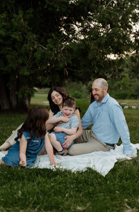 Mother holding her son while daughter laughs and looks at their dad during a family photoshoot in the grass in Maine — candid New England family photography