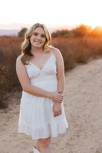 Smiling high school senior girl standing on a dirt path at sunset wearing a white dress, senior portrait session by Kaitlyn Dawn Photography.