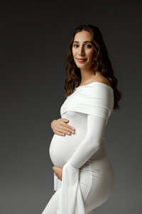 Pregnant woman in an elegant white gown smiling softly while cradling her baby bump during a maternity photoshoot in Yucaipa by Kaitlyn Dawn Photography.