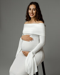 Smiling pregnant mom seated on a stool in a white gown, holding her belly during a maternity session by Kaitlyn Dawn Photography in the Inland Empire.