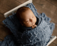 Close-up portrait of a sleeping newborn baby wrapped in textured blue fabric and posed on a rustic wooden floor during a professional newborn photography session by Kaitlyn Dawn Photography.
