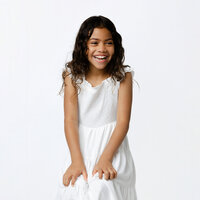 Young girl in a white dress sitting against a white studio backdrop, smiling naturally during a classic child portrait session by Kaitlyn Dawn Photography, professional children’s photographer.