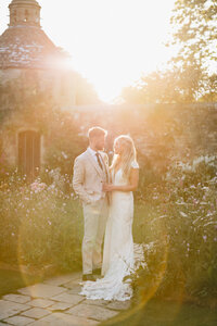 A couple stands on a garden path at Nymans National Trust during sunset. The man is in a light beige suit, and the woman wears a white lace gown. They are surrounded by flowers, with a stone building in the background, bathed in warm, golden light.