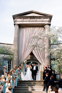 Bride and groom raising their hands in celebration after being pronounced married during their ceremony in front of the grand columned entrance at Burritt on the Mountain in Huntsville, Alabama, photographed by Perry Hancock.