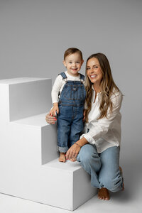 Mother sitting on white studio steps holding her toddler son, relaxed and joyful studio family portrait captured by Kaitlyn Dawn Photography, modern neutral family photography studio.