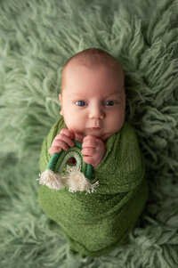 Newborn baby with bright blue eyes swaddled in green, holding a tiny rainbow toy on a fluffy green blanket at a Waukesha, Wisconsin photography studio.