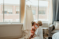 Bride kisses young daughter while getting ready on wedding day, wedding dress hanging in window background.