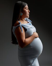 Pregnant woman in a light blue maternity dress posing in profile during a dramatic studio maternity portrait by Kaitlyn Dawn Photography.