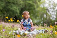 Jacksonville, FL portrait session capturing Baby boy in denim overalls sitting on a blanket among wildflowers with a yellow flower tucked behind his ear, gazing upward with wide-eyed curiosity during an outdoor child milestone session in Palatka.