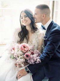 groom whispering to bride as she smiles with eyes closed holding a lush pink and lilac bouquet