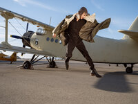 Person in a brown coat caught mid-movement beside a white biplane on an airfield tarmac.