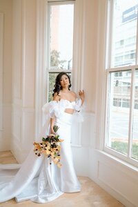 Capturing the elegance as a model poses in a wedding gown by a sunlit window, complemented by a vibrant bouquet for an impactful styled photoshoot.