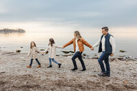 A family of four, holding hands, strolls along a rocky beach with calm water in the background. Captured by a Toronto Family Photographer, they're dressed in casual fall clothing under a cloudy sky.