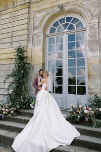 Bride in a flowing white gown and veil embracing groom on stone steps, framed by floral arrangements and tall arched doors of an elegant estate.