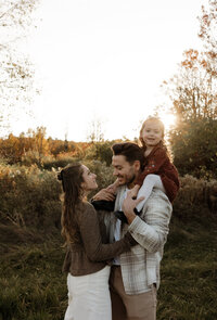 A golden-hour family photo capturing dad carrying his daughter on his shoulders while mom smiles up at them. Warm, honest, and intimate lifestyle photography in Maine.