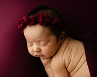 Sleeping newborn baby girl wearing a burgundy floral headband posed on a deep red backdrop during a studio newborn photography session by Yucaipa newborn photographer Kaitlyn Dawn Photography.