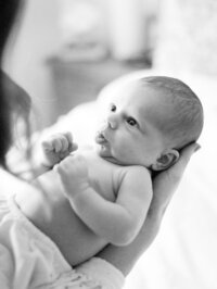Black and white close-up of an awake newborn baby looking up while being cradled in mom’s arms during an in-home lifestyle session.